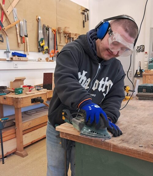 Ein Mann mit Handschuhen, Gehörschutz und Schutzbrille fertigt ein Werkstück aus Holz