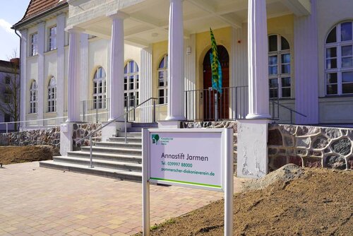 Ein Schild mit der Gebäudebezeichnung steht vor dem Annastift Jarmen, einem Gebäude mit Treppe, Säulen und großen alten Fenstern.