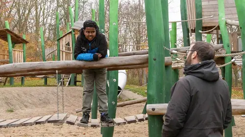 Ein Junge steht auf einem Klettergerüst und wird von einem Mann am Rand angeschaut